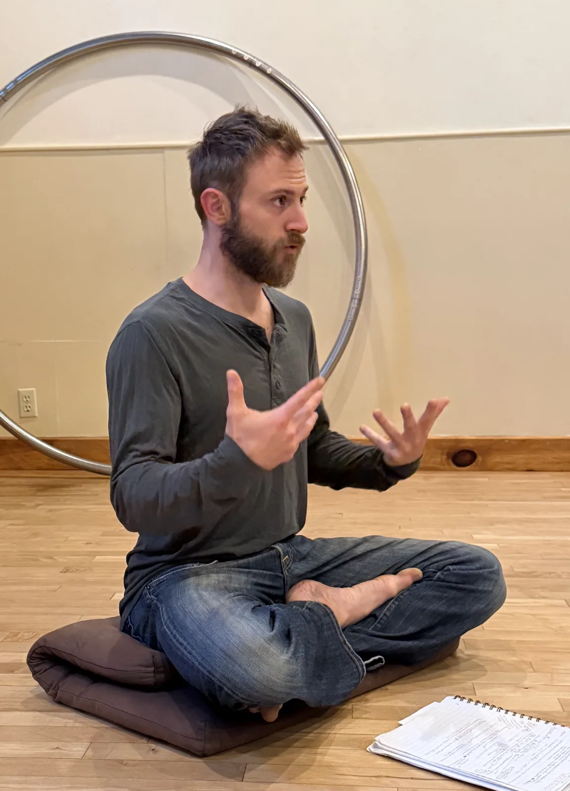 John sitting cross-legged on a cushion on a wooden floor, speaking with open hands, a large metal hoop behind him.