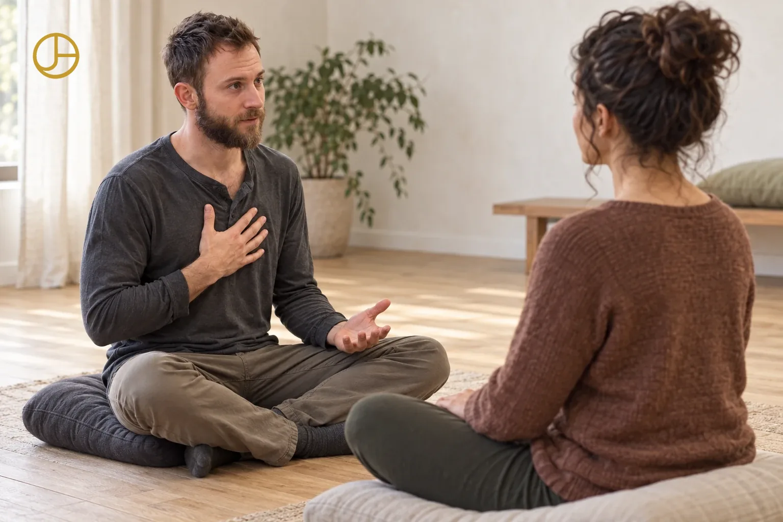 John sits cross-legged on a floor cushion, one hand on his chest, speaking with a woman seated across from him.